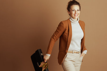 A cheerful businesswoman in a brown blazer and beige stands confidently with her hand in her pocket. She holds a briefcase with autumn leaves, projecting a modern, professional, and successful imageの写真素材