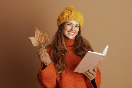 A cheerful young woman in a yellow beanie and orange sweater smiles at the camera, holding an autumn leaf and an open book. This cozy image captures a joyful fall mood.の写真素材