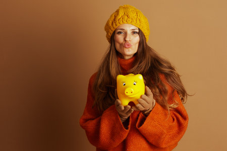 A playful woman in a cozy orange sweater and yellow knit hat puckers her lips, holding a yellow piggy bank. The soft lighting highlights her thoughtful and whimsical approach to financial savings.の写真素材