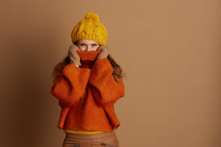 Playful woman in knit hat and oversized orange turtleneck pulls sweater collar up with gloved hands, partially hiding face while looking at camera. This image captures cozy autumn or winter warmthの写真素材
