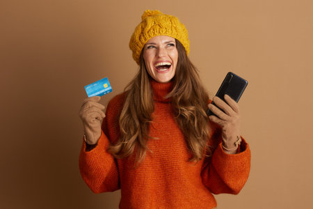 A joyful woman in a warm orange sweater and yellow hat laughs while holding a credit card and smartphone, symbolizing the ease and pleasure of online purchasing.の写真素材