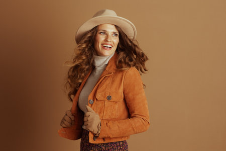 Beaming woman showcases stylish autumn ensemble, featuring light tan fedora, orange suede jacket, and patterned skirt. Joyful expression captures essence of fall fashion against warm brown background.の写真素材