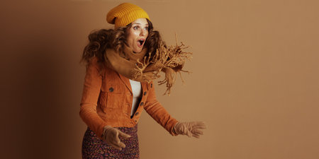 A surprised woman with curly hair, wearing a yellow beanie, tan scarf, and rust-colored jacket, is captured mid-motion, wide-eyed with an open mouth and outstretched hands.の写真素材