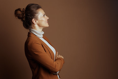 A middle-aged businesswoman with light brown hair in a bun stands in profile, gently smiling with arms crossed, wearing a rust-brown blazer and light turtleneck against a solid background.の写真素材