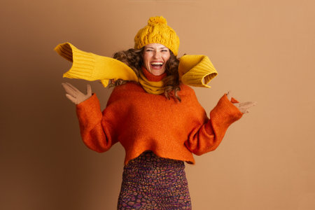 A joyful woman with curly brown hair is captured mid-jump, arms outstretched, scarf billowing. She wears a yellow beanie, scarf, orange sweater, and patterned skirt against a light brown background.の写真素材