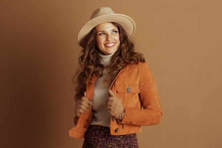 A confident woman with curly brown hair, wearing a beige fedora, turtleneck, and an orange suede jacket, poses against a plain background. Her stylish autumn ensemble is captured in a medium shot.の写真素材