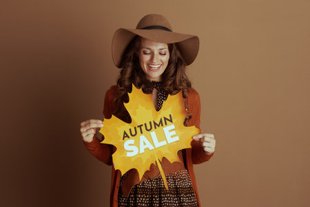 A cheerful woman in a wide-brimmed hat and orange cardigan holds an "AUTUMN SALE" sign, looking down happily, promoting seasonal discounts.の写真素材