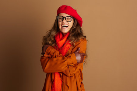 A cheerful young woman in a red beret, black glasses, and a warm brown coat laughs with eyes closed and arms crossed, embodying pure joy and cozy autumn comfort.の写真素材