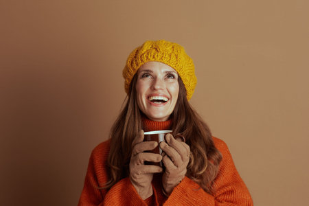 A smiling woman with long brown hair, wearing yellow knitted hat, orange turtleneck sweater, and...
