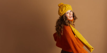 A laughing woman with curly brown hair wears a bright yellow pom-pom hat, orange knit sweater, and...
