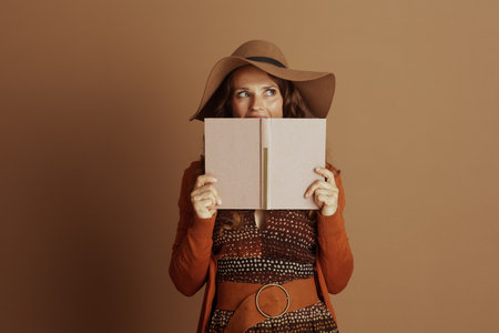 A woman in an autumn-inspired outfit, featuring a wide-brimmed hat and orange cardigan, holds a pink book to her face with a curious expression against a muted brown backdrop.の写真素材