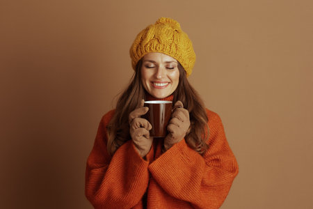 An adult woman in yellow knit hat and orange sweater holds brown mug with both hands, smiling with closed eyes. Against tan background, she embodies warmth, comfort, and contented autumn self-careの写真素材