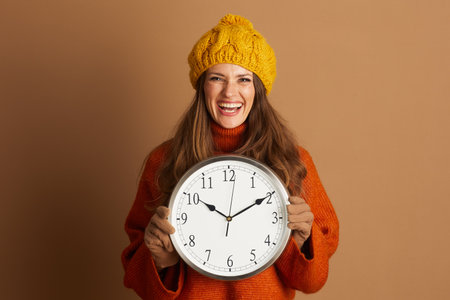 A smiling woman in a yellow knitted hat and orange turtleneck holds a large analog clock, looking at the camera. This cheerful image suggests autumn time changes and seasonal preparation.の写真素材