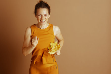 A woman in orange activewear smiles while jogging, holding yellow autumn leaves. This energetic...
