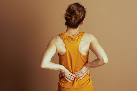 A woman in a vibrant yellow-orange athletic top stands with her hands pressing into her lower back,...