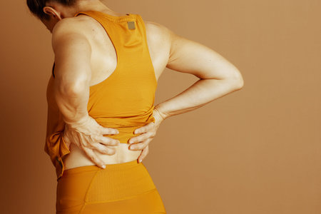 A woman, seen from behind, clutches her lower back in pain while wearing mustard yellow athletic...