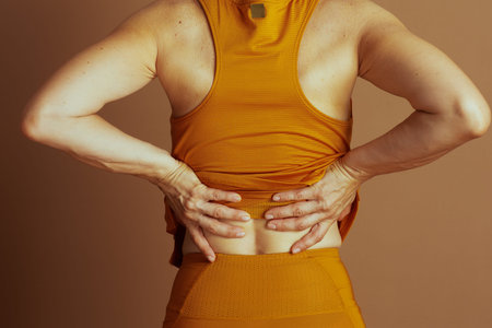 An adult, seen from behind, clutches their lower back, indicating discomfort or pain. They wear an...