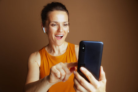 A smiling woman in an orange top, wearing earbuds, looks at her smartphone and points at the screen....