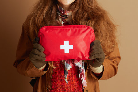 Person in warm outdoor gear, including brown coat and red sweater, holds red first aid kit with white cross. This medium shot emphasizes safety and preparedness for outdoor adventures or travel.の写真素材