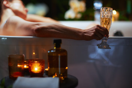 A person relaxes in a white bathtub, enjoying a sparkling beverage in a patterned glass. Lit candles and warm lighting create a luxurious, tranquil atmosphere for self-care and indulgence.の写真素材