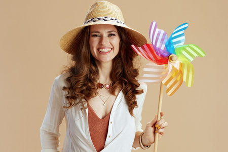 Beach vacation. smiling modern 40 years old housewife in white blouse and shorts against beige background with windmill toy and straw hat.の写真素材