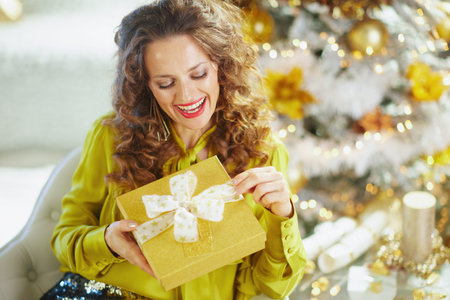 A woman with curly brown hair and bright smile joyfully opens glittery gold gift box with...