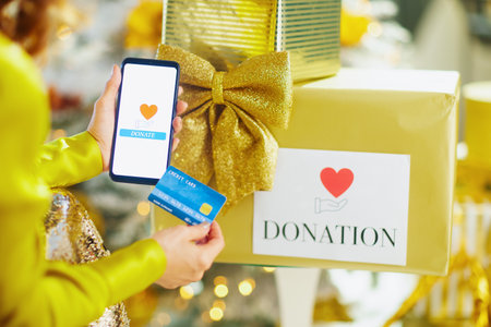 A close-up shows hands holding smartphone displaying donation app and credit card over gold gift box. The festive background of blurred lights highlights theme of charitable giving during holidaysの写真素材