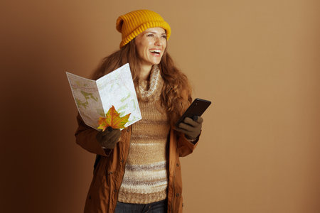 A smiling woman, clad in yellow knit hat and brown coat, holds map and smartphone while looking up, possibly envisioning journey. This medium shot captures spirit of autumn travel and explorationの写真素材