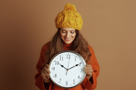 A smiling woman in a yellow beanie and orange turtleneck sweater holds a large wall clock, looking down at it. Her warm, cheerful expression against a brown background evokes a cozy autumn mood.の写真素材