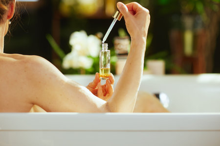 A woman, seen from behind, dispenses yellow bath liquid via dropper into a white bathtub. Serene...