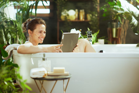 A smiling woman finds peace in a white bathtub, using her digital tablet amidst a vibrant indoor...