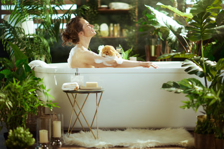 A woman, smiling with eyes closed, finds serenity in a bubbly bath, cleansing with a natural sponge...