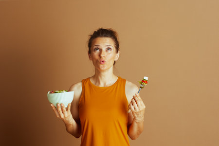 A woman in an orange top, with a thoughtful expression and pursed lips, looks upward while holding a bowl of salad.の写真素材