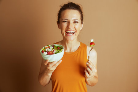 A smiling woman in an orange tank top, hair in a bun, holds a salad with a fork, featuring tomato and mozzarella. This medium shot against a soft brown background conveys a positive, healthy lifestyleの写真素材