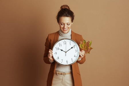 A smiling woman in professional attire gently holds a large clock and autumn leaves, embodying...