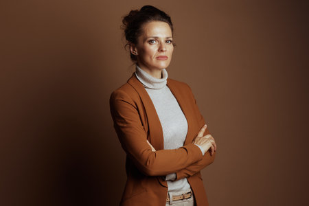 A poised businesswoman with dark hair and a serious, confident expression stands with arms crossed, wearing a brown blazer and beige turtleneck against a solid brown studio background.の写真素材