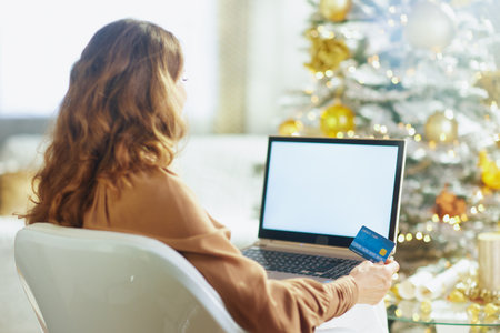 A woman with long brown hair sits in a white chair, engrossed in online shopping with a credit card or laptop. A festive Christmas tree with golden ornaments creates a cozy, soft-lit holiday mood.の写真素材