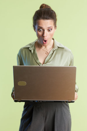 A surprised woman looks down at her laptop in shock or excitement, wearing a green blouse and gray...