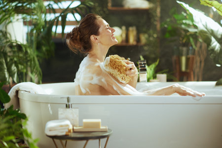 A woman with closed eyes & a serene smile enjoys a bubble bath, washing her shoulder with a natural...