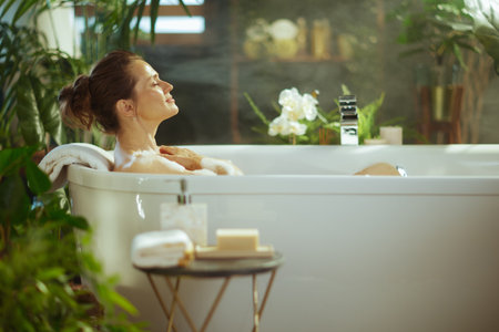A serene woman with eyes closed and a peaceful smile relaxes in a white bubble bath, hands on her chest. The modern bathroom, with lush green plants & soft steam, creates a tranquil self-care oasis.の写真素材