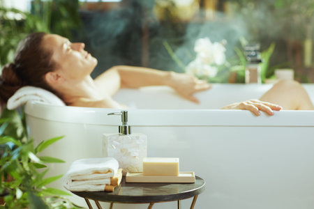 An adult woman with eyes closed reclines in a steaming white bathtub, enveloped in tranquility....
