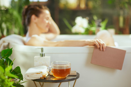 An adult woman, eyes closed, relaxes peacefully in a white bathtub, a book in hand. A tea mug & towels are arranged. Soft natural light illuminates green plants in this tranquil bathroom oasis.の写真素材