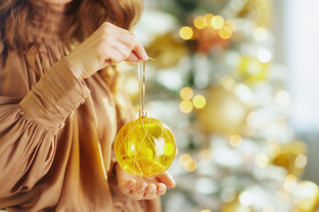 An elegant woman in a brown dress delicately holds a shimmering golden Christmas ornament. A blurred Christmas tree with warm, golden bokeh lights forms a festive and soft background.の写真素材