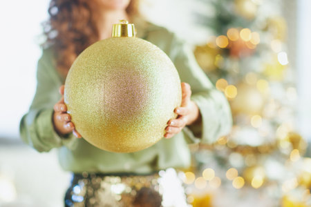 An elegant woman with curly brown hair, dressed in a silky olive green top and sequined trousers, holds a glittering gold Christmas ornament amidst soft, festive lighting.の写真素材