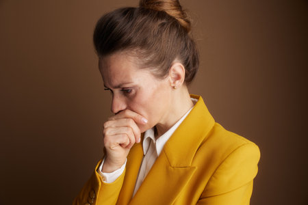 A professional woman in a yellow blazer, seen in profile, holds her hand to her mouth with a worried or thoughtful expression, contemplating a difficult business decision.の写真素材
