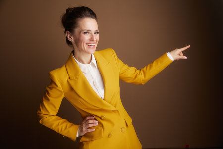 A smiling businesswoman in a yellow blazer extends her arm, elegantly pointing rightward to offer guided insight or highlight an important detail in a professional presentation.の写真素材