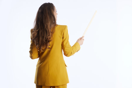 A professional woman in a mustard yellow suit, seen from the back, holds a wooden pointer stick, symbolizing instruction, presentation, or guiding an audience in a bright studio.の写真素材