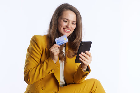 A confident businesswoman in a bright yellow suit smiles while holding a credit card and smartphone, embodying modern online finance and secure digital transactions.の写真素材