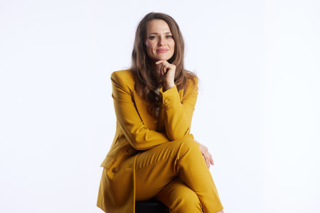 A confident businesswoman in a vibrant yellow pantsuit poses seated against a clean white studio background. Her hand rests thoughtfully on her chin, conveying professionalism and strategic thinking.の写真素材