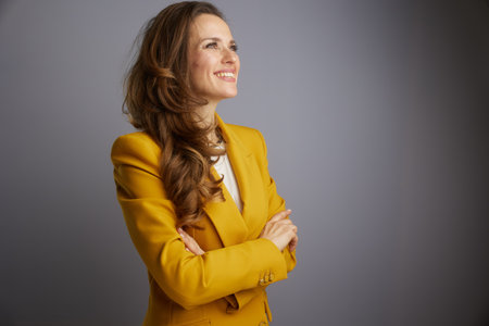A professional woman with long, wavy brown hair, wearing a bright yellow blazer, stands in confident profile with arms crossed against a gray background, exuding joy.の写真素材